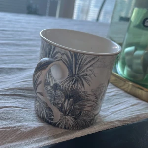 Vit mugg med växtmotiv - En stilren vit mugg med ett detaljerat svart växtmotiv. Perfekt för morgonkaffet eller teet. Muggen har en klassisk form med ett bekvämt handtag.